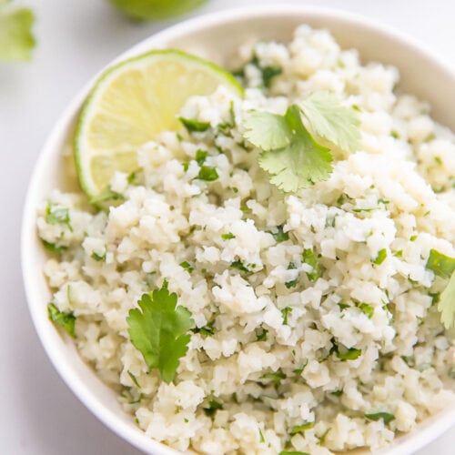 Cilantro lime cauliflower rice, garnished with a lime wedge, in a white bowl on a marble countertop.