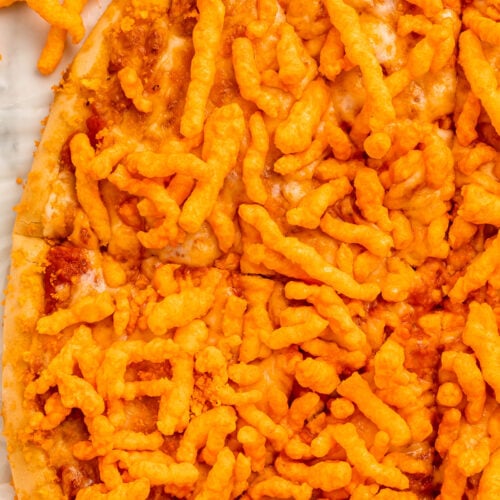 A large round cheese pizza topped with bright orange crunchy Cheetos, cut into triangle-shaped slices but still together as a whole pizza.