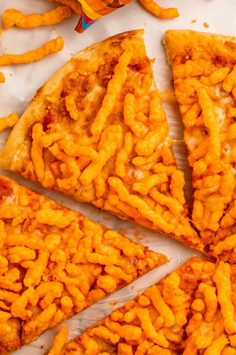 A slice of cheese pizza topped with crunchy Cheetos, being pulled away from other slices of pizza.