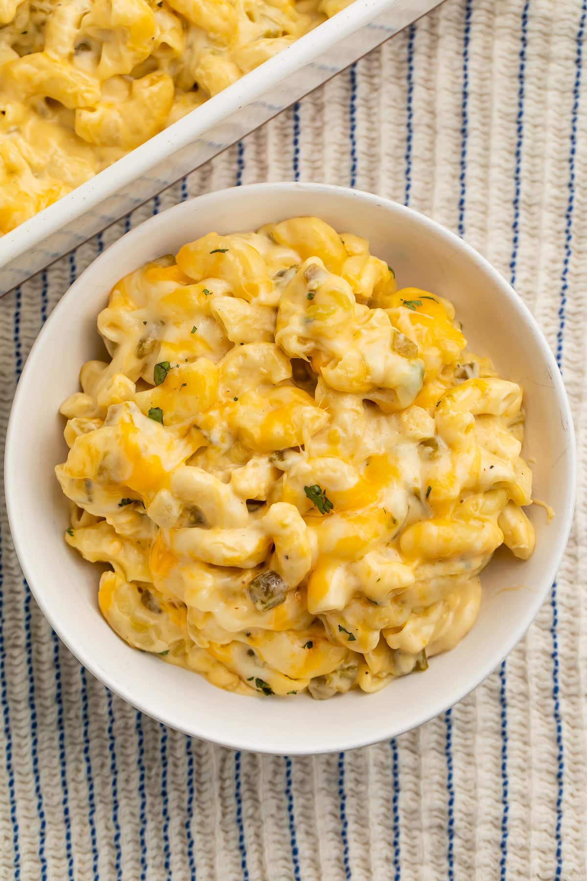 Green chili mac and cheese in a white bowl next to a white casserole dish of mac and cheese on a table.