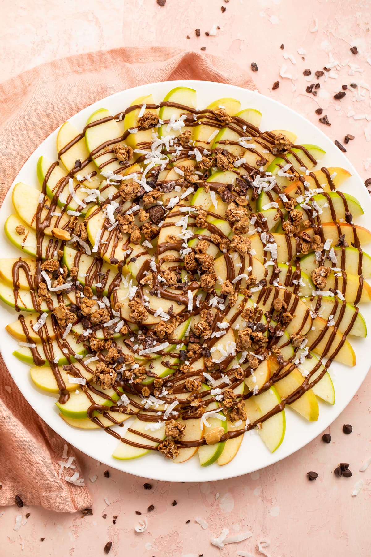 Overhead, top-down look at a plate of green and red apple slices topped with swirls of chocolate syrup and granola.