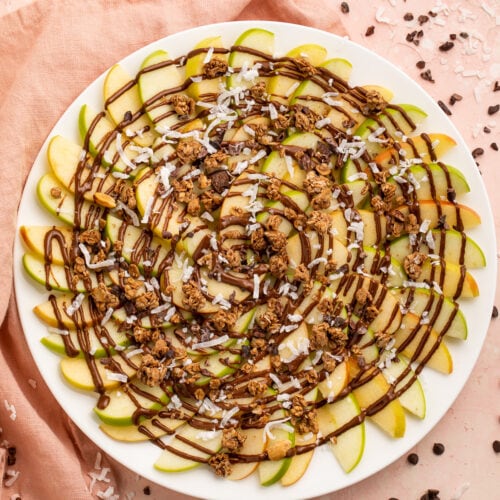 Overhead, top-down look at a plate of green and red apple slices topped with swirls of chocolate syrup and granola.