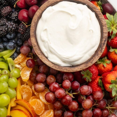 A small wooden bowl containing a white, whipped cream cheese dip on a platter surrounded by a rainbow of fruit.