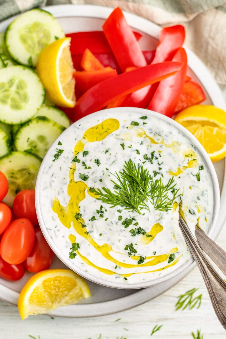 A bowl of creamy Whole30 tzatziki dotted with green fresh herbs and topped with a drizzle of olive oil, surrounded by cucumber slices, grape tomatoes, and bell pepper slices.