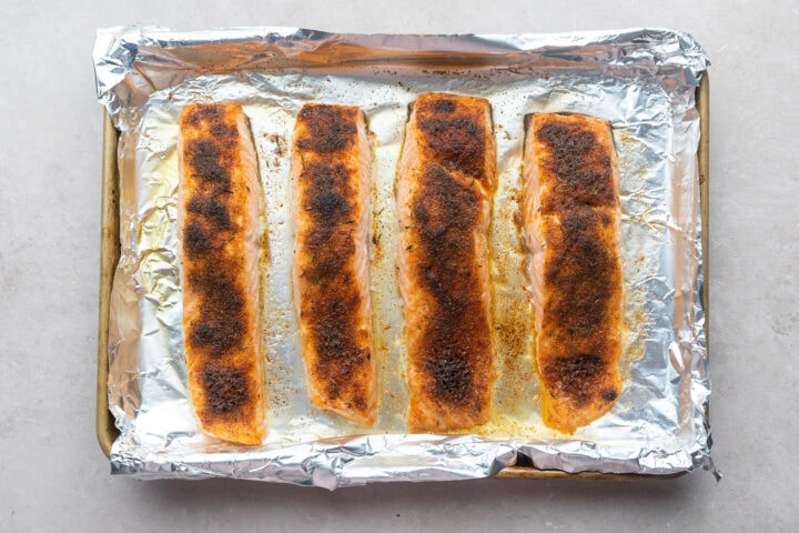 Four salmon fillets on a foil-lined baking sheet, topped with neutral oil and jerk seasoning.