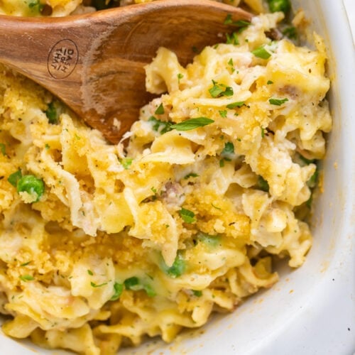 A large oval casserole dish holding tuna noodle casserole, with a large wooden spoon resting in the dish.