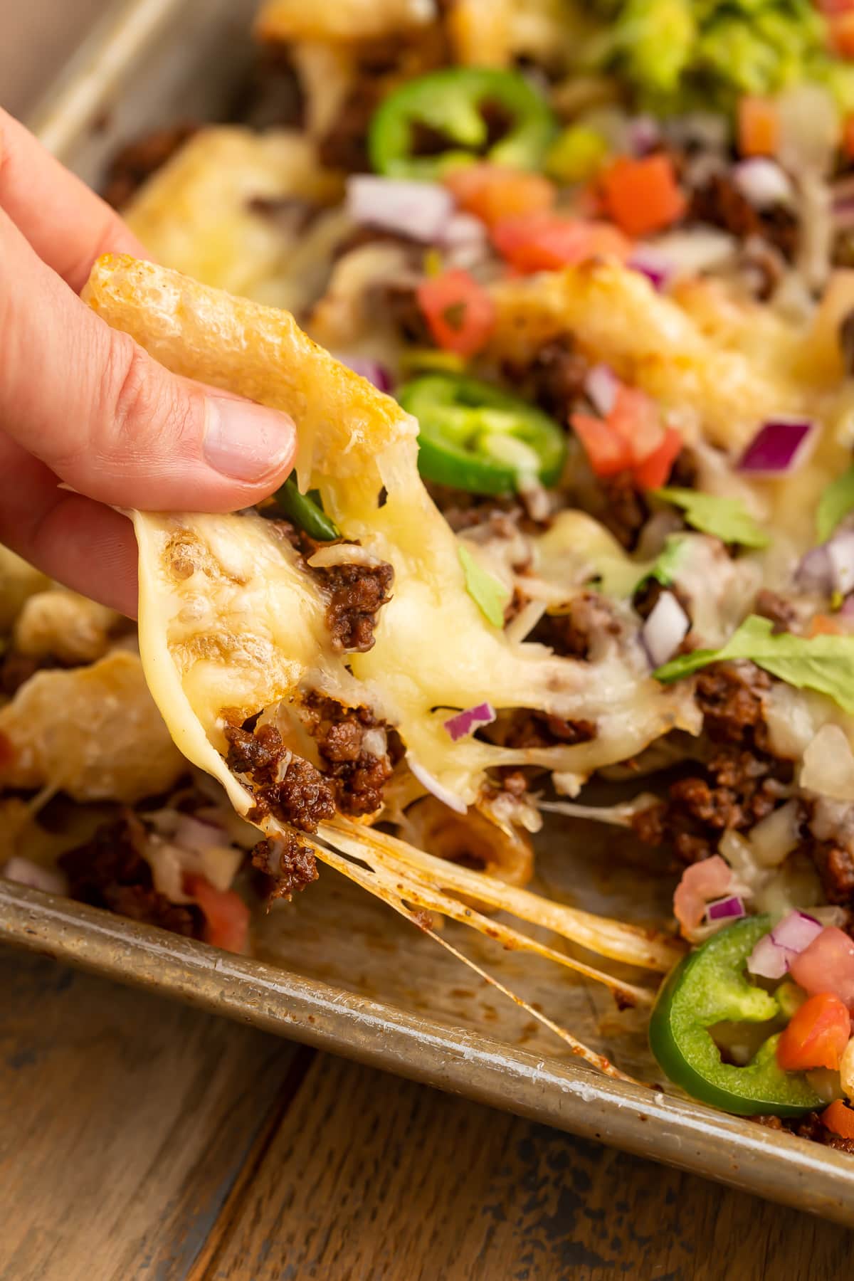 A white woman's hand using a pork rind as a tortilla chip to scoop up nacho toppings and melted cheese.