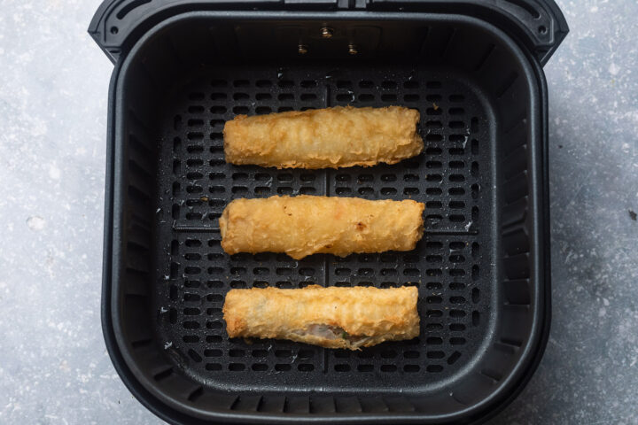 Three frozen egg rolls in a black air fryer basket.