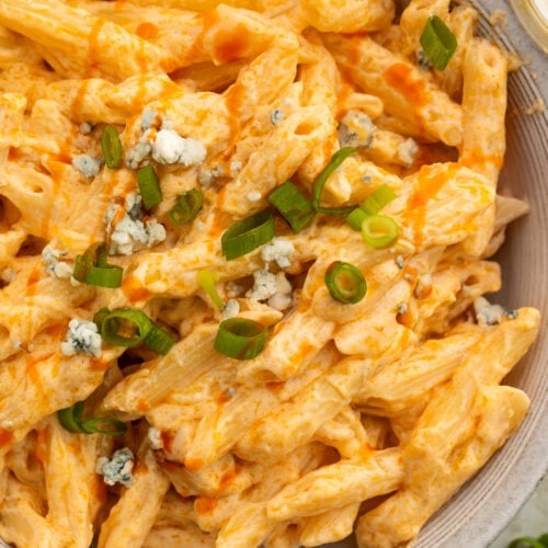 Close-up of an off-white bowl holding creamy buffalo chicken alfredo made with penne pasta and topped with sliced green onions.