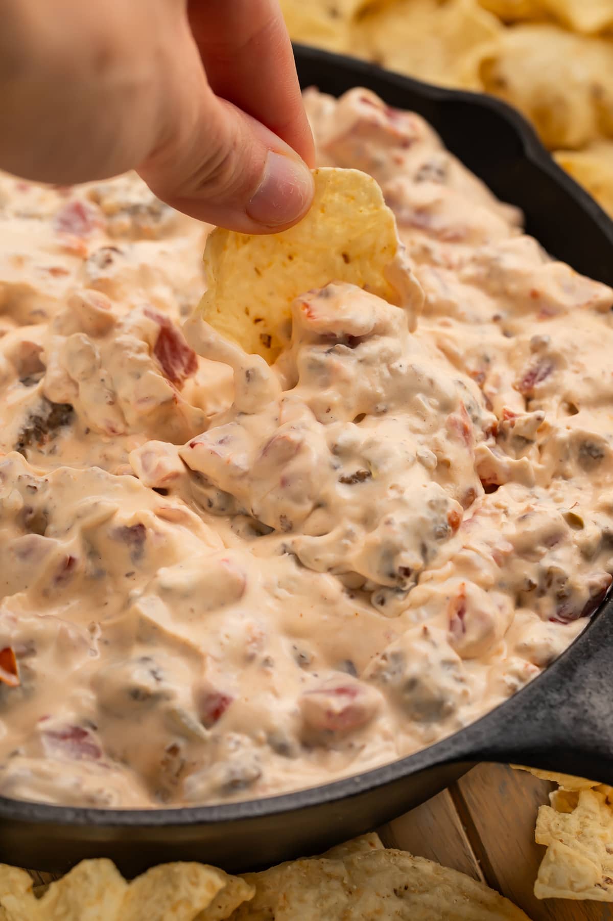 A white woman's hand dipping a scoop-shaped tortilla chip into a skillet full of cream cheese sausage dip.