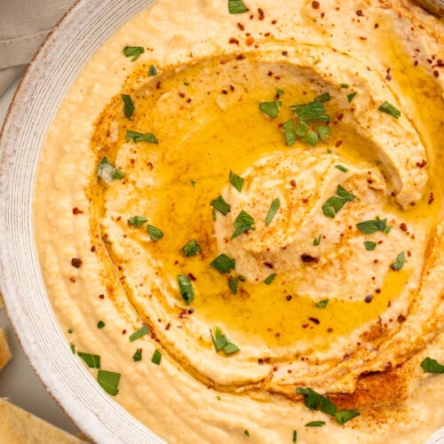 Close-up overhead view of a white bowl holding hummus made without tahini, swirled with olive oil and garnished with herbs.