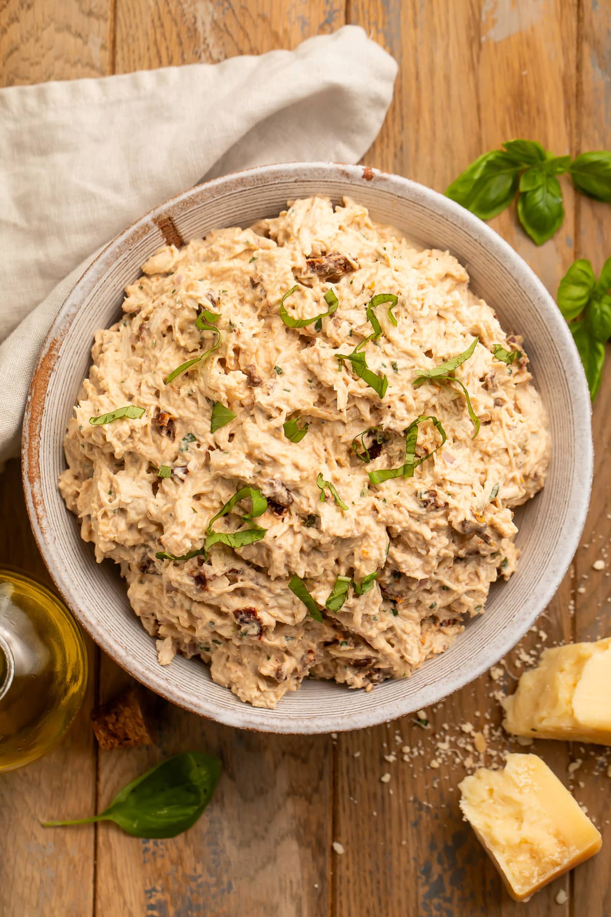 A zoomed-out look of a bowl of marry me chicken salad on a stained wood tabletop with a neutral cloth napkin.