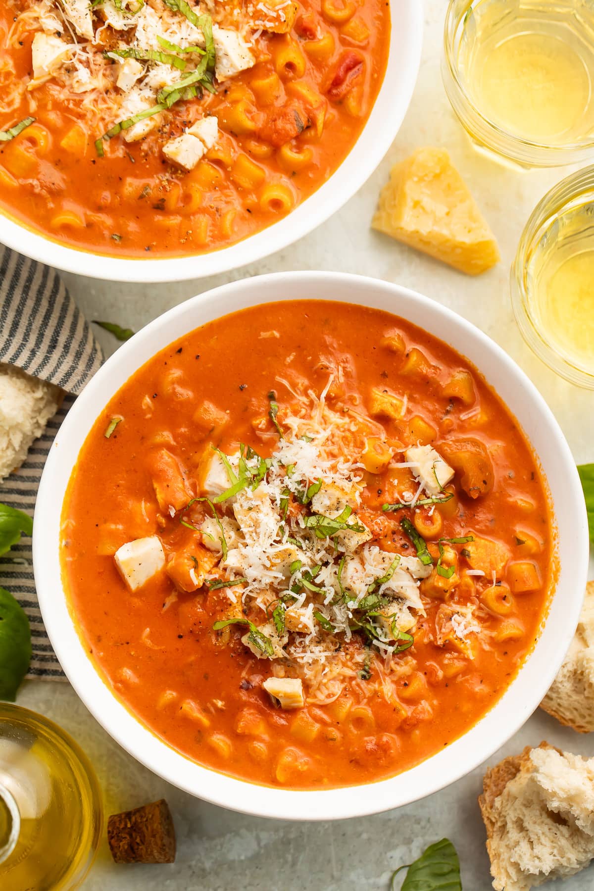 Overhead view of two white soup bowls holding deep red-orange tomatoey soup with ditalini pasta topped with diced chicken, shredded parmesan, and thin strips of basil.