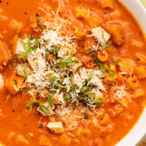 Close-up of a white soup bowl holding deep red-orange tomatoey soup with ditalini pasta topped with diced chicken, shredded parmesan, and thin strips of basil.