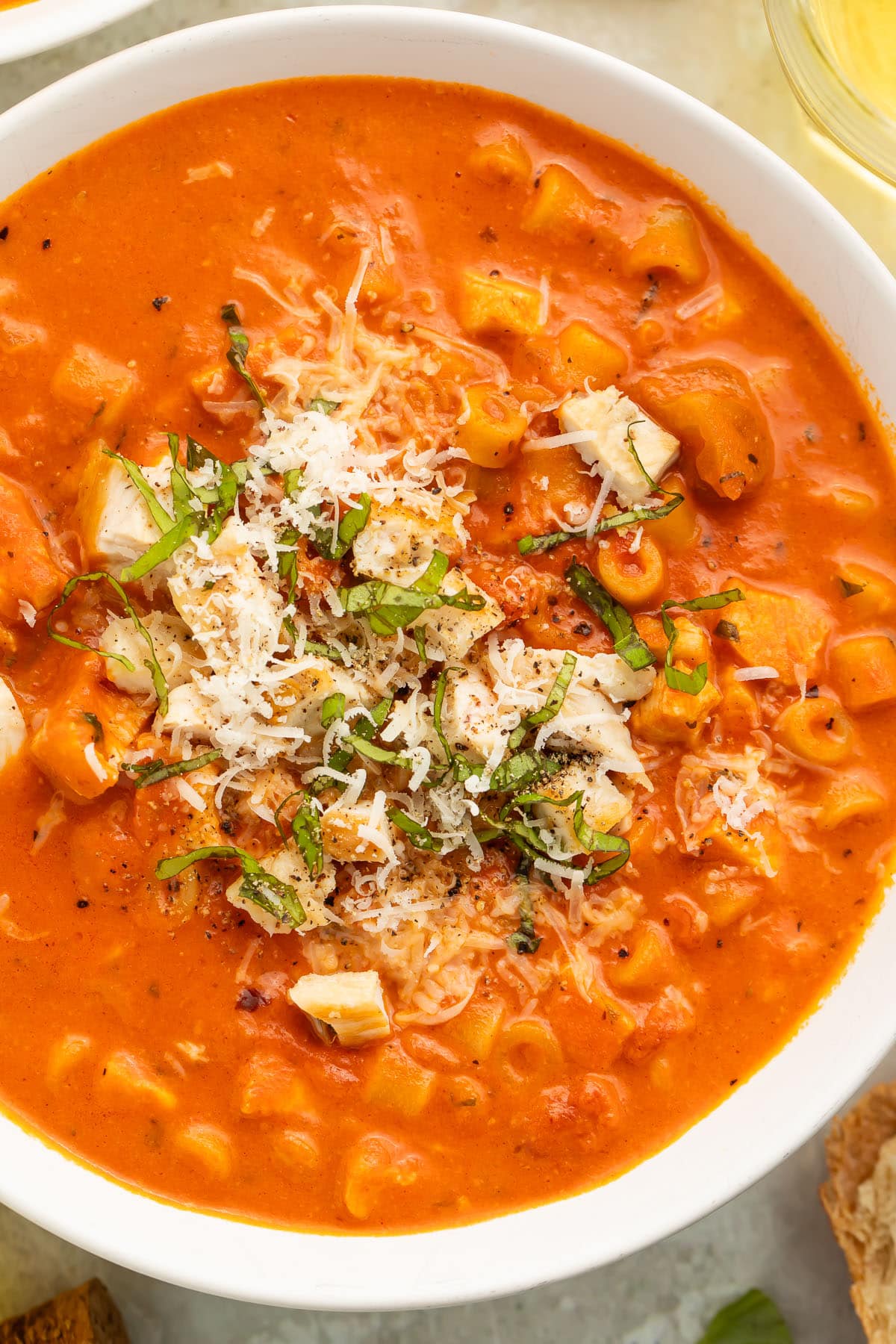 Close-up of a white soup bowl holding deep red-orange tomatoey soup with ditalini pasta topped with diced chicken, shredded parmesan, and thin strips of basil.