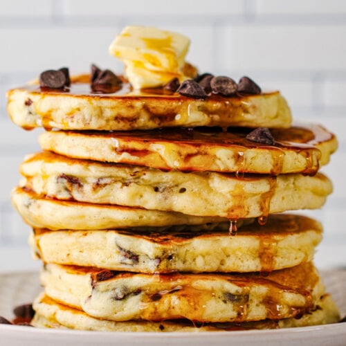 A stack of chocolate chip pancakes topped with chocolate chips, two pats of butter, and plenty of maple syrup.