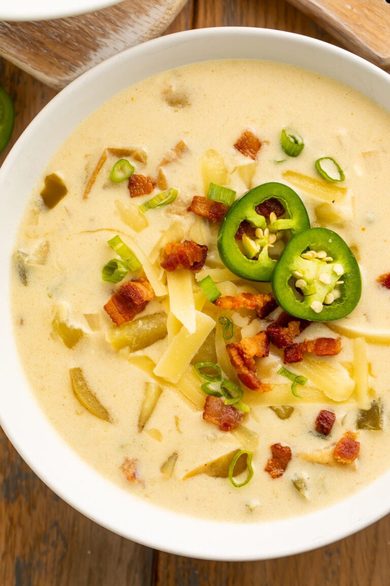 Top-down view of a large white bowl holding pale yellow, creamy jalapeño soup, topped with sliced jalapeños, diced bacon, and shredded cheese.