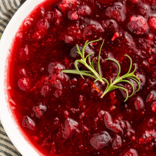 Close-up of whole berry rosemary cranberry sauce in a white bowl with a garnish of fresh rosemary.