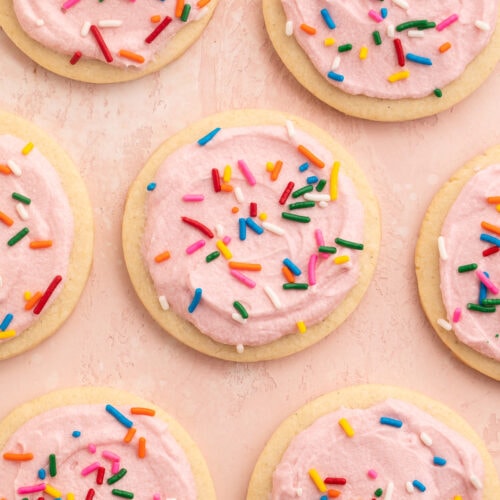 Rows of gluten free sugar cookies topped with pink buttercream frosting and rainbow jimmies on parchment paper.