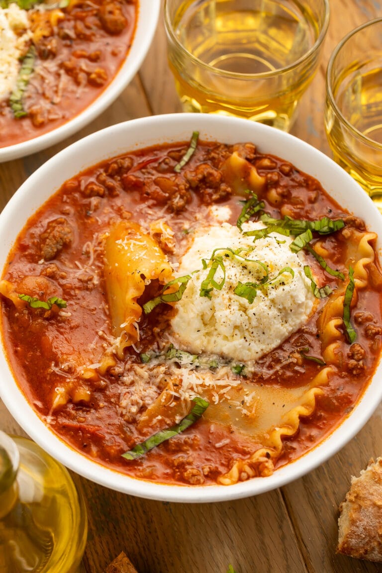 A bowl of lasagna soup on a wooden table next to a piece of bread and a glass of wine.