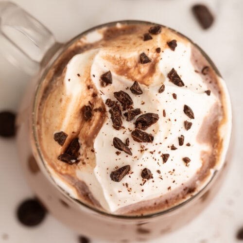 Top-down photo of a glass coffee mug holding deep, rich paleo hot chocolate topped with whipped coconut cream and chocolate shavings.