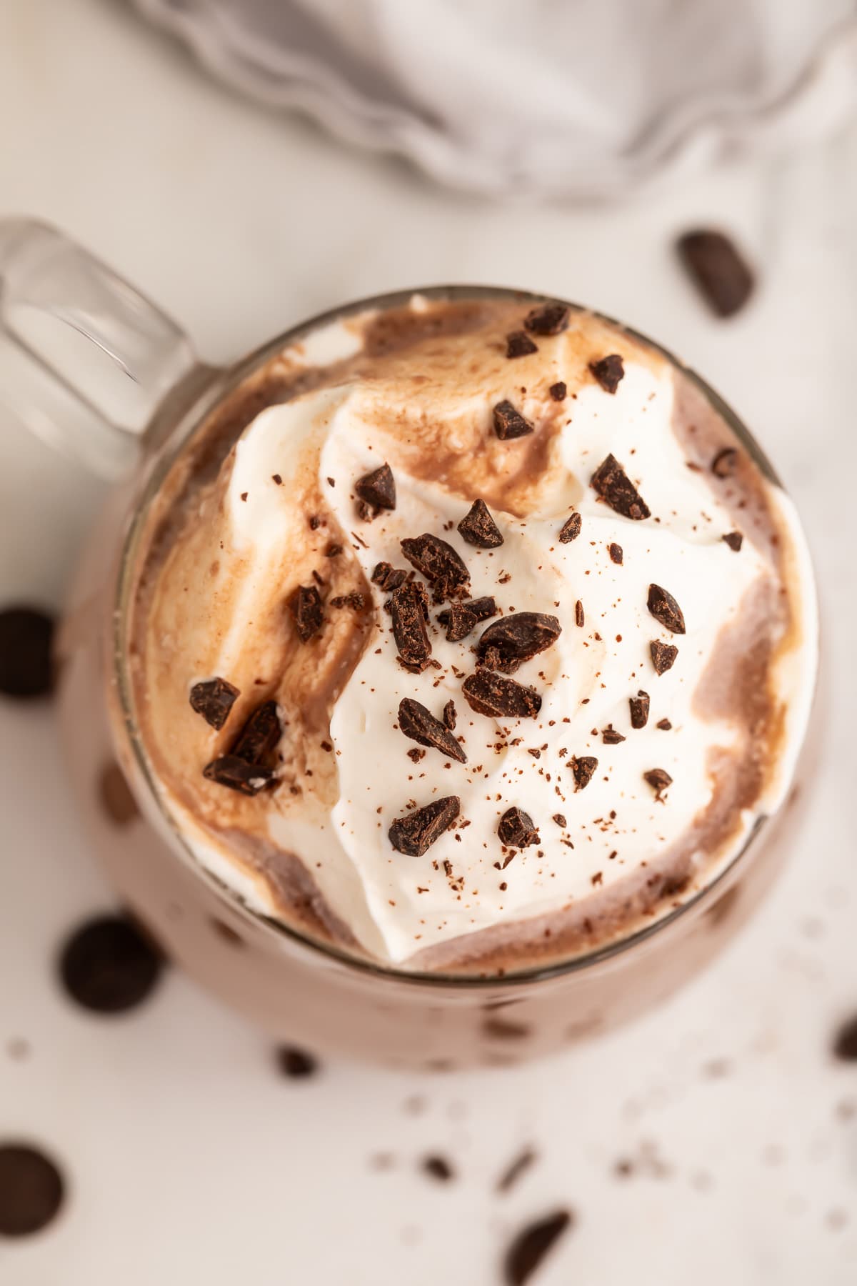 Top-down photo of a glass coffee mug holding deep, rich paleo hot chocolate topped with whipped coconut cream and chocolate shavings.