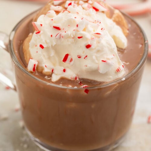 A clear glass mug of rich paleo peppermint hot chocolate topped with whipped coconut cream and candy cane pieces.