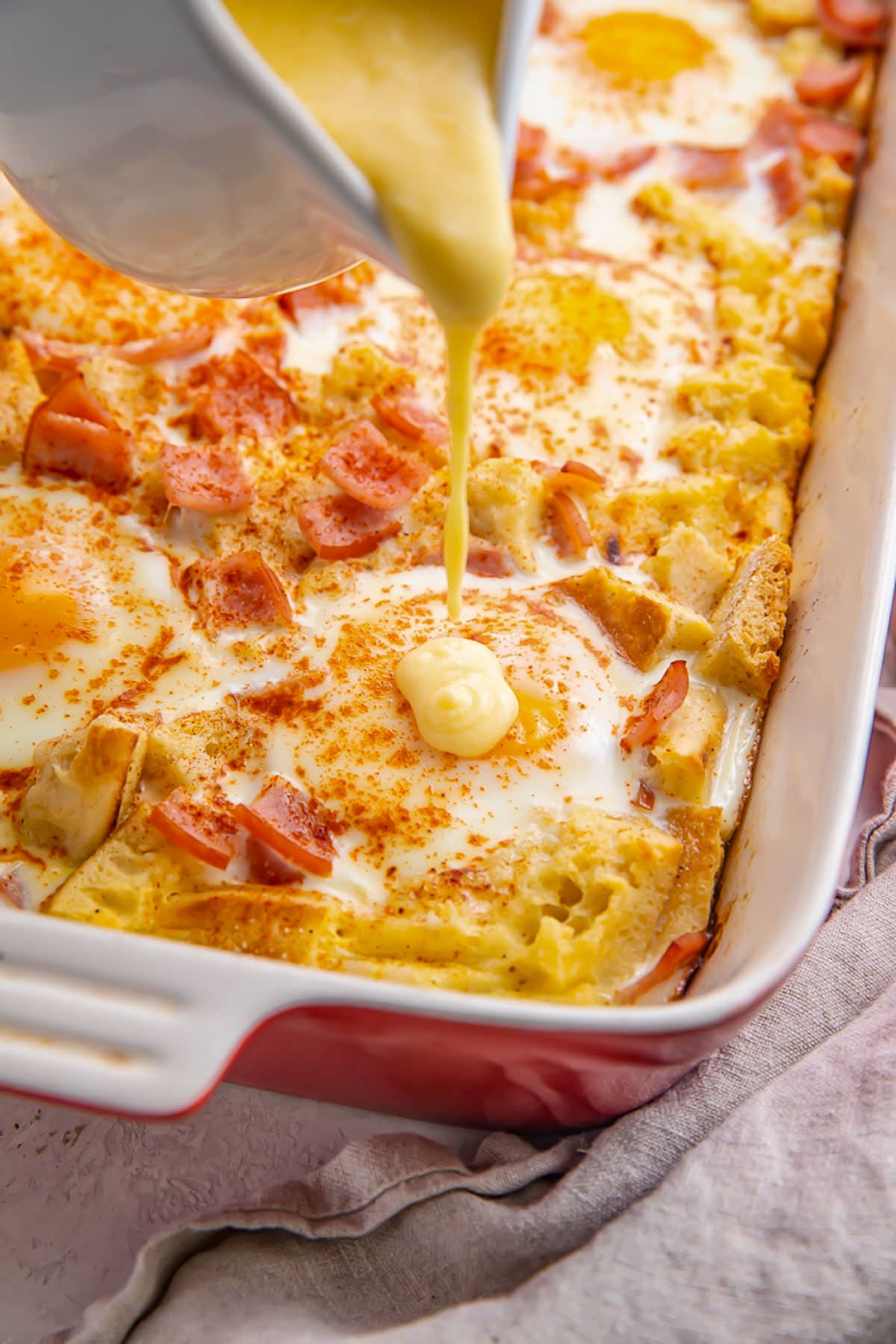 Creamy hollandaise sauce being poured over an eggs benedict casserole in a casserole dish.