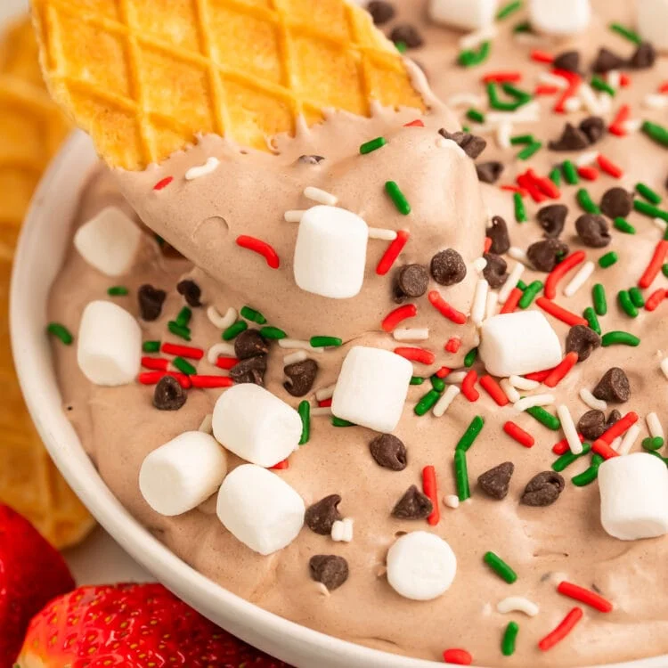 A woman's hand holding a waffle cookie being used to scoop hot cocoa dip out a large bowl.