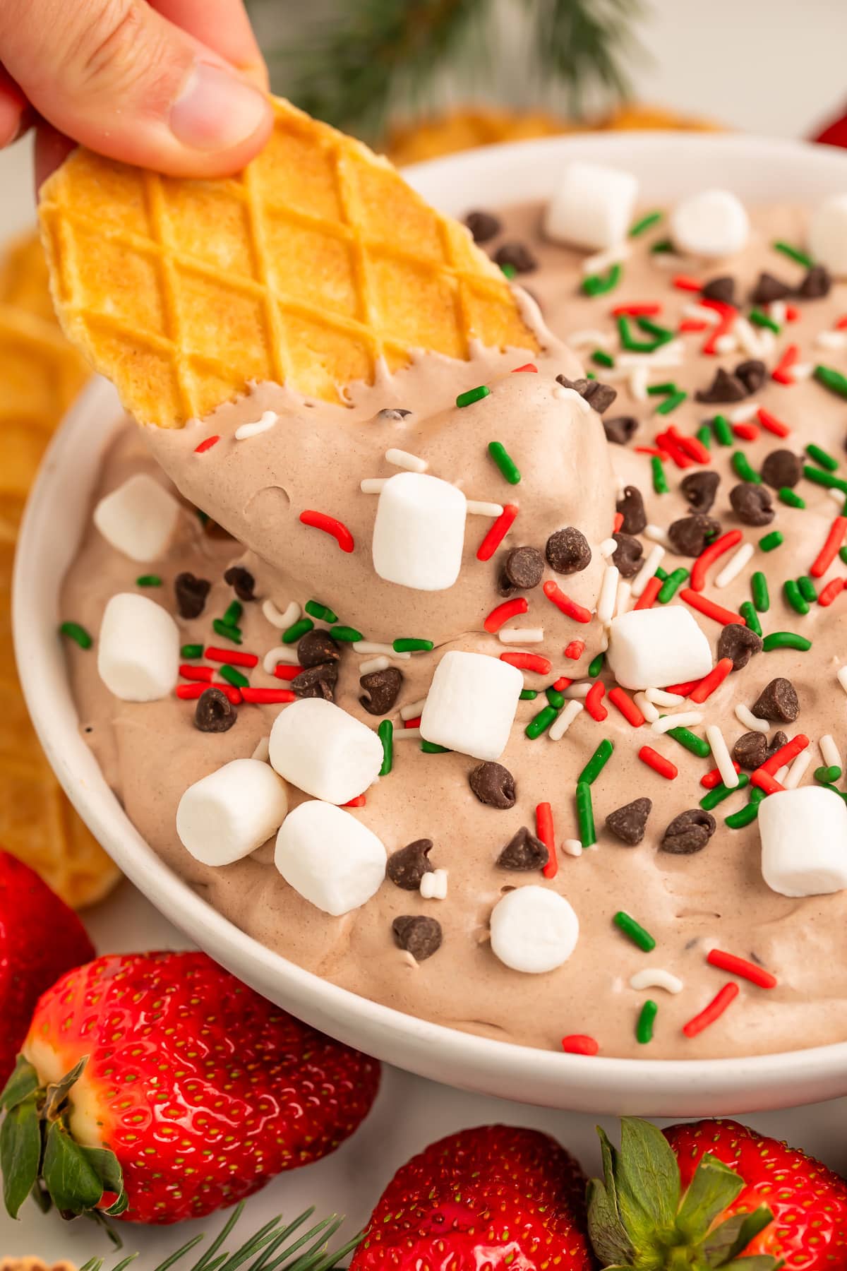 A woman's hand holding a waffle cookie being used to scoop hot cocoa dip out a large bowl.