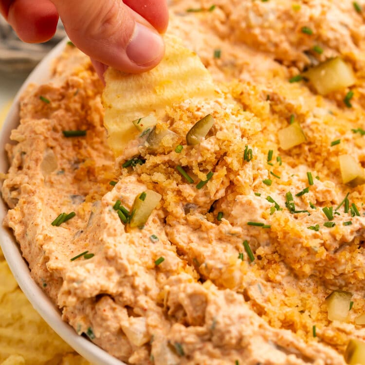 A white woman's hand holds a ridged potato chip, using it to scoop Nashville hot chicken dip out of a large bowl.