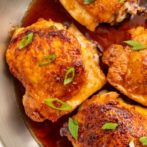 A close-up of bone-in, skin-on chicken thighs in a large silver skillet with a hot honey sauce.