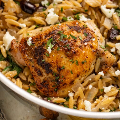 Close-up of a Greek seasoned chicken thigh in a skillet with orzo and feta.