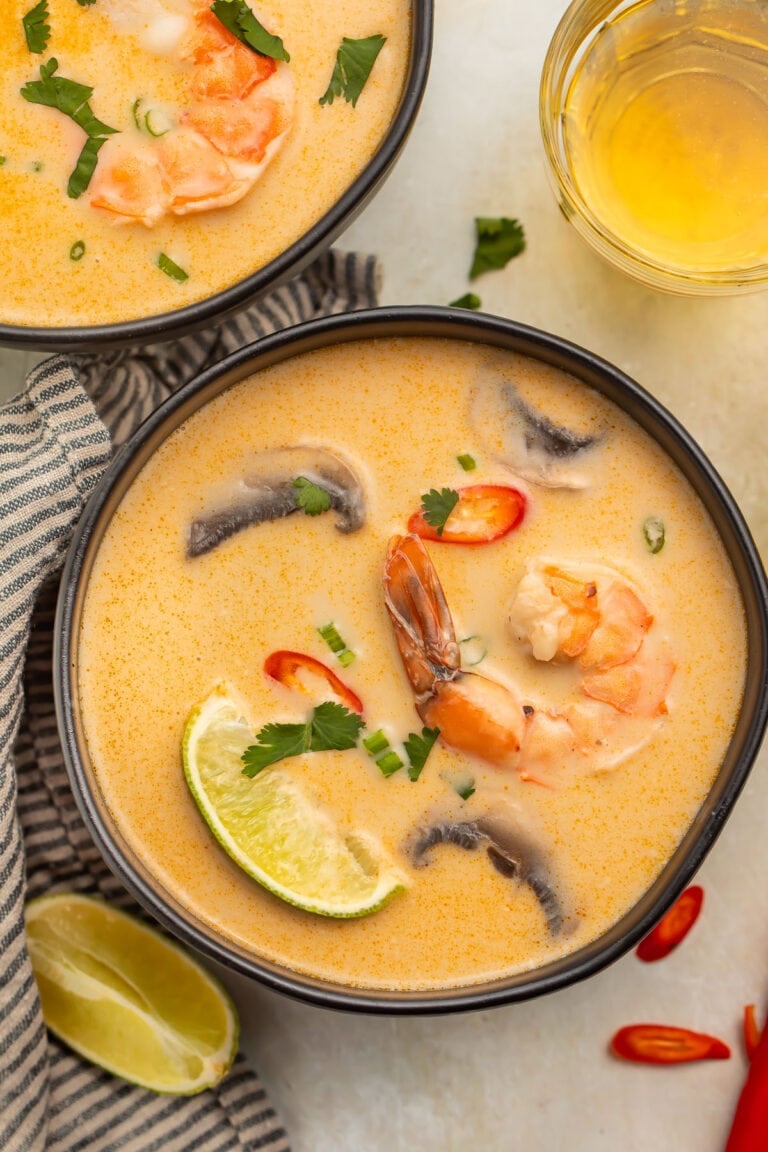 An overhead photo of a black soup bowl holding tom kha goong (Thai coconut shrimp soup) with lime wedges, shrimp, and aromatics floating in the broth.