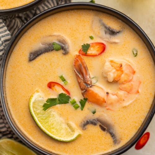 An overhead photo of a black soup bowl holding tom kha goong (Thai coconut shrimp soup) with lime wedges, shrimp, and aromatics floating in the broth.