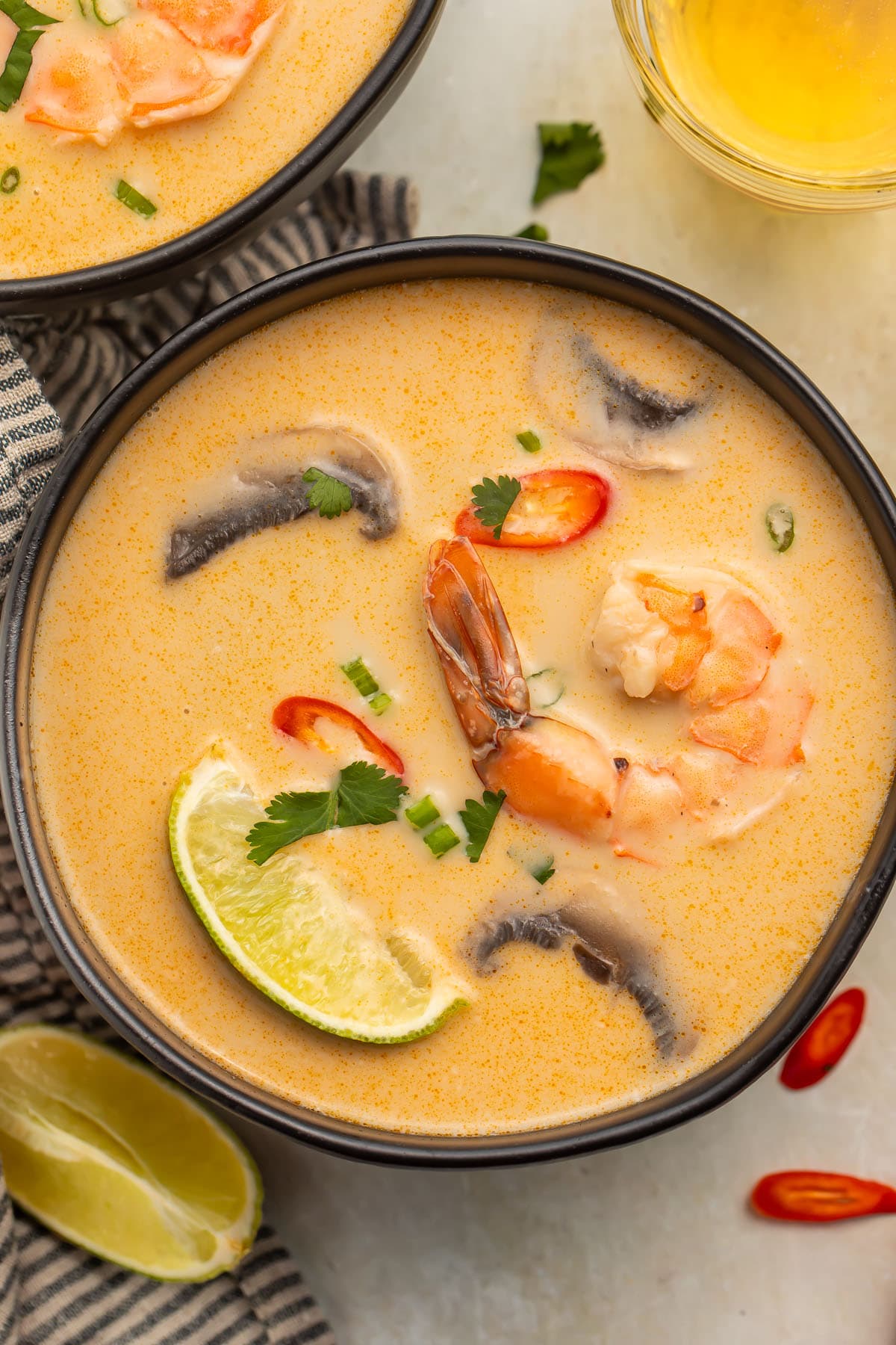 An overhead photo of a black soup bowl holding tom kha goong (Thai coconut shrimp soup) with lime wedges, shrimp, and aromatics floating in the broth.