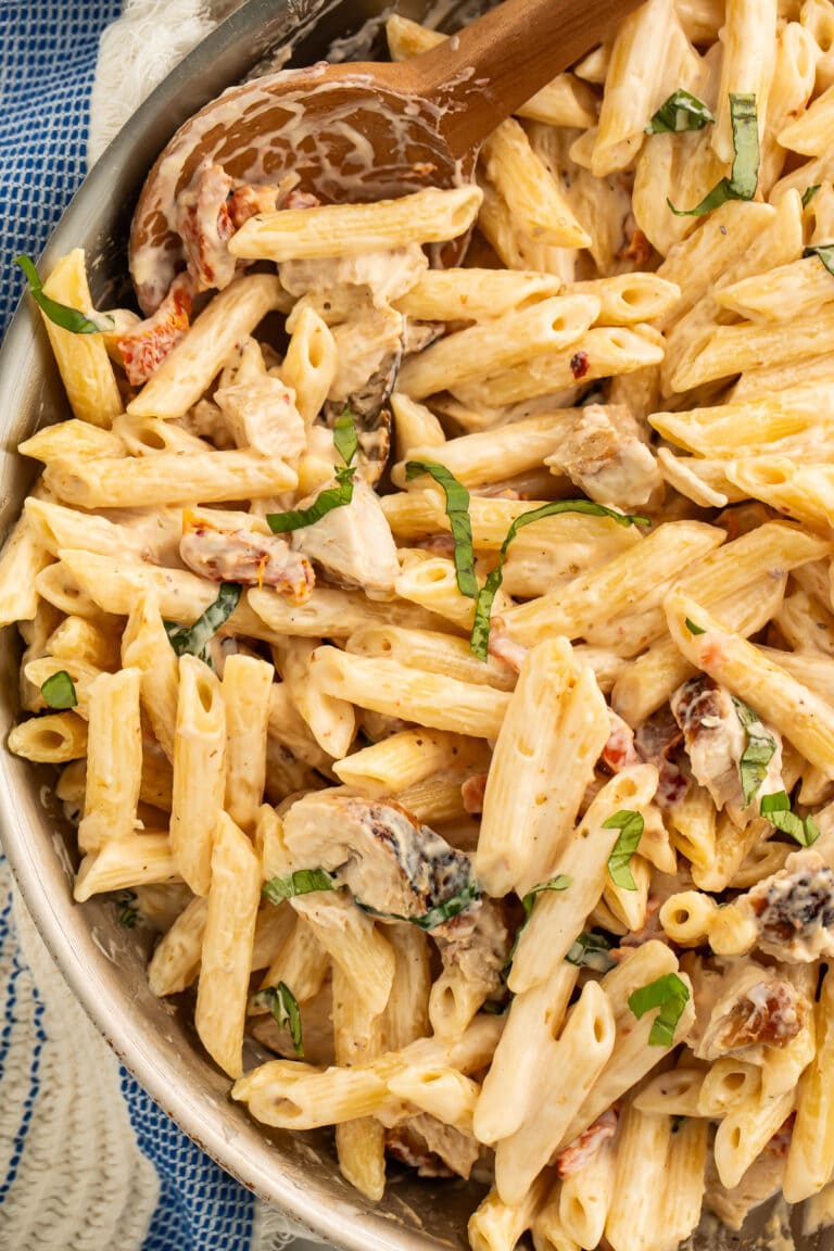 A large silver skillet of penne pasta in a sun-dried tomato cream sauce with chicken and basil.