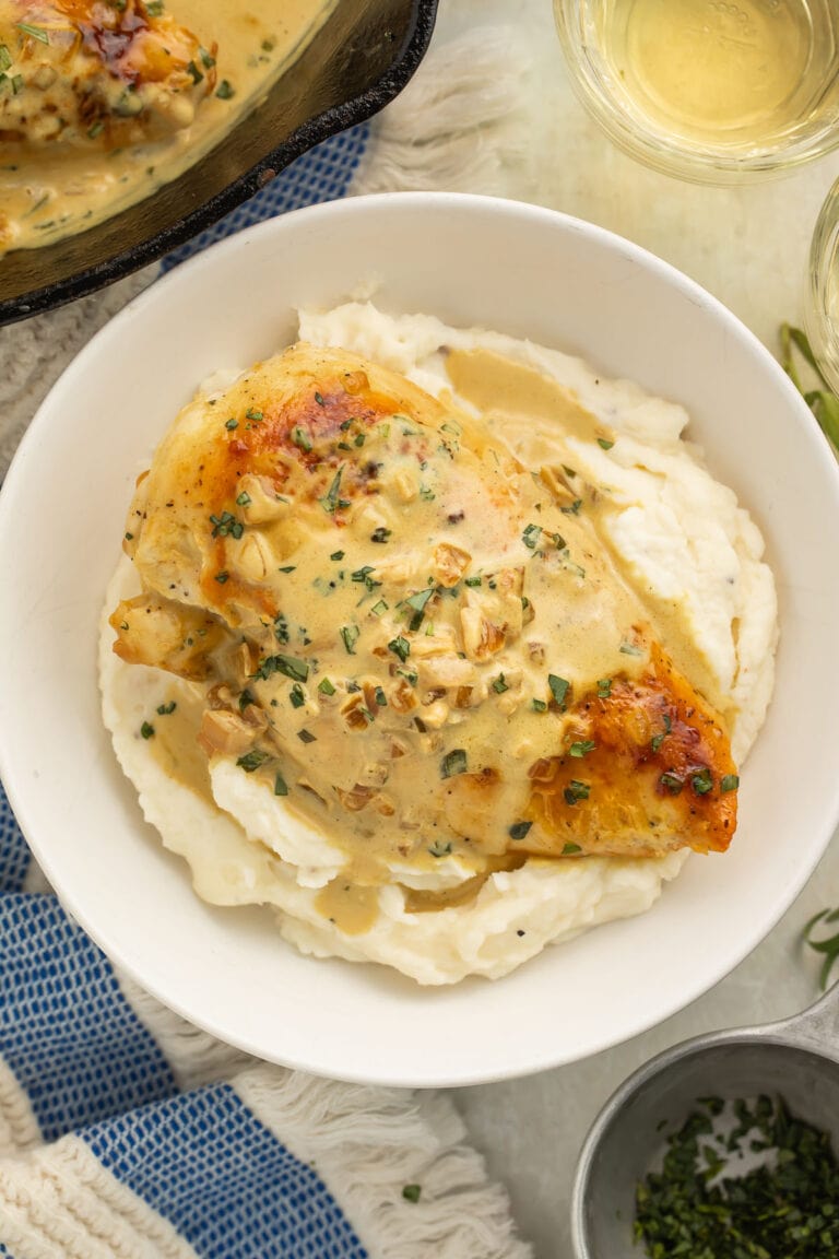 Creamy dijon chicken breast placed on top of a bed of mashed potatoes in a large white bowl on a table with a blue and white stripe dish towel.