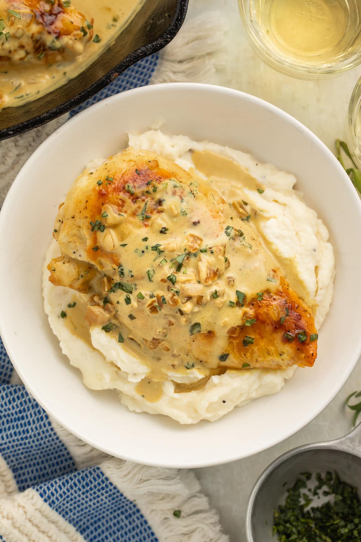 Creamy dijon chicken breast placed on top of a bed of mashed potatoes in a large white bowl on a table with a blue and white stripe dish towel.