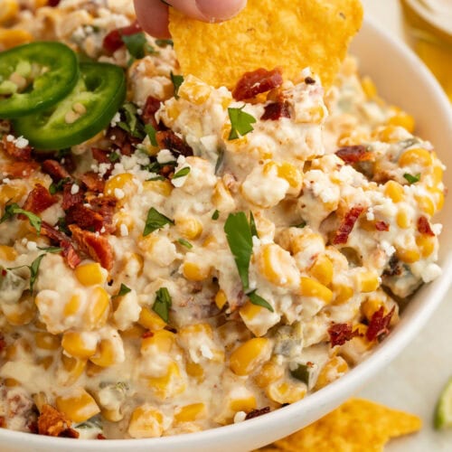 A white woman's hand holding a tortilla chip that's being used to scoop jalapeño corn dip out of a large bowl.