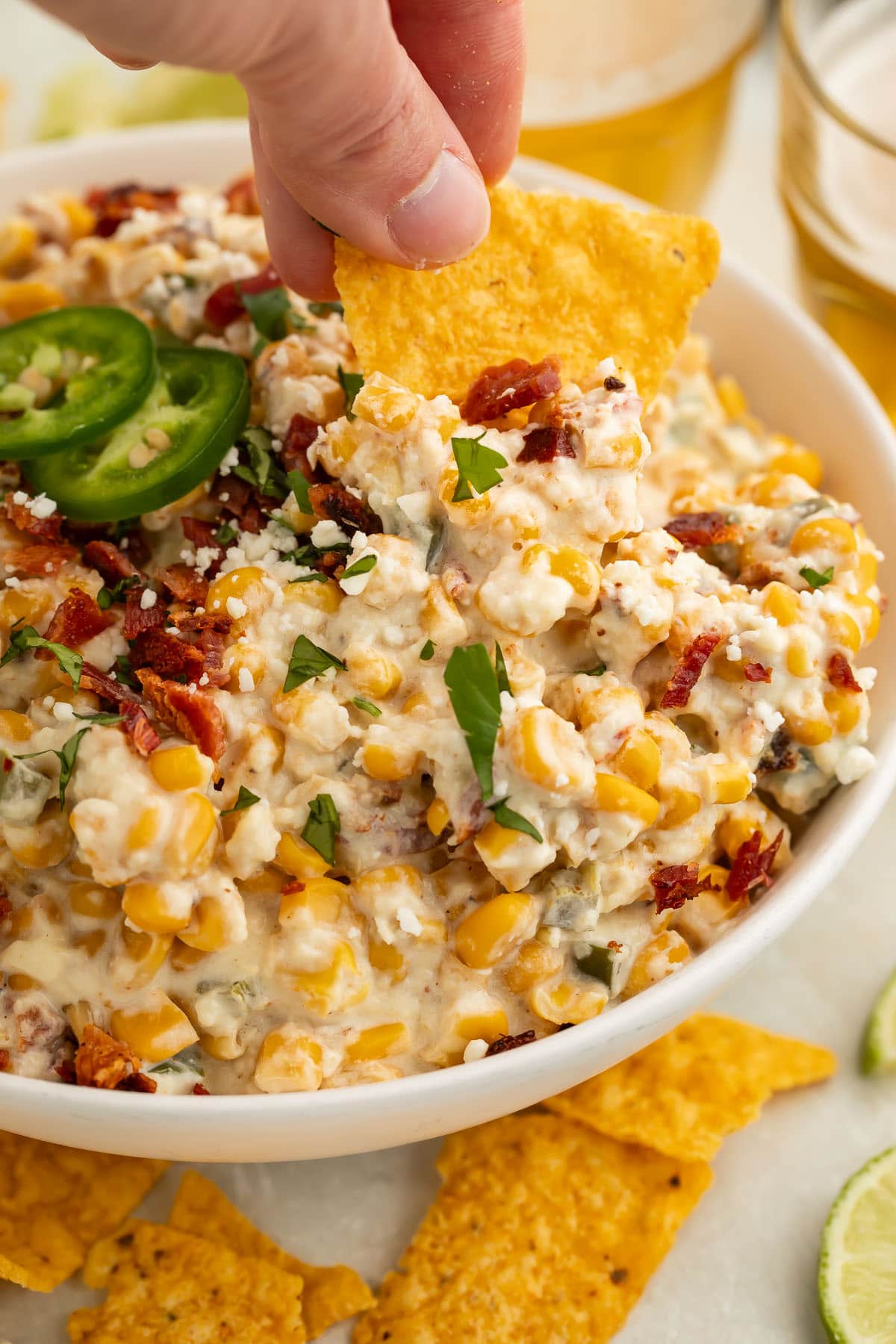 A white woman's hand holding a tortilla chip that's being used to scoop jalapeño corn dip out of a large bowl.