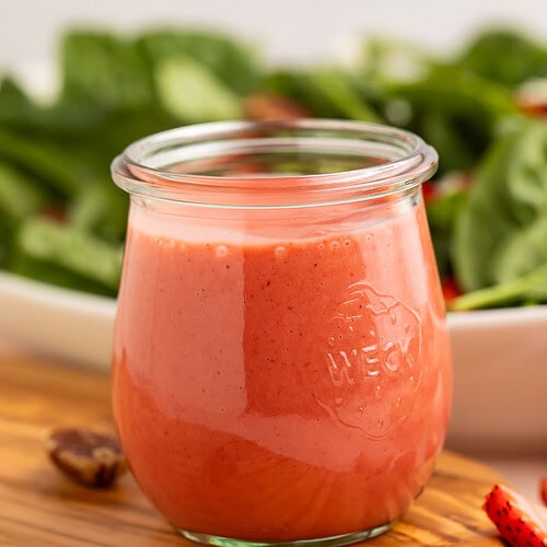 A jar of creamy pale red strawberry vinaigrette in front of a leafy spinach salad.