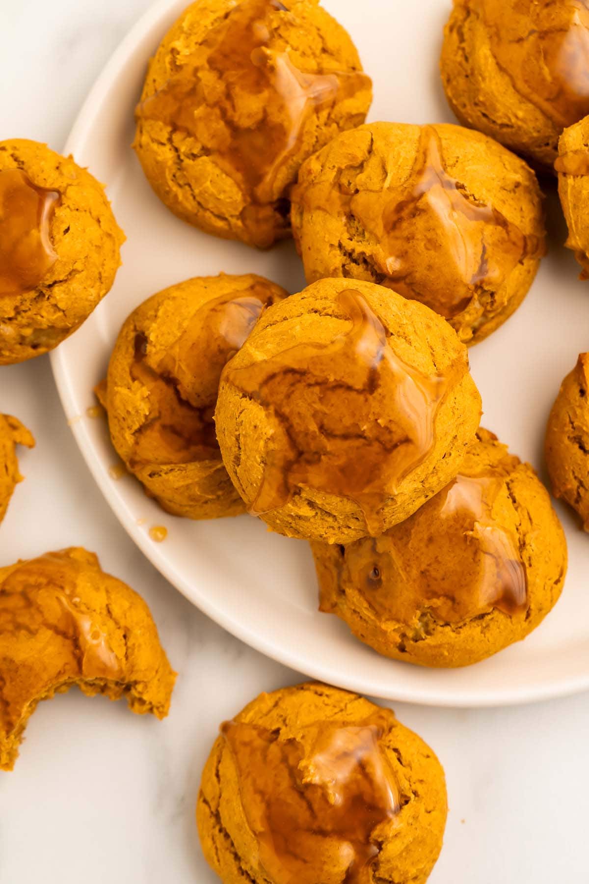 Pumpkin cookies topped with a light maple glaze arranged on and around a round white plate on a table.