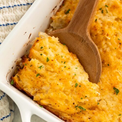 A large wooden spoon resting in a square casserole dish holding a cheesy potato casserole.