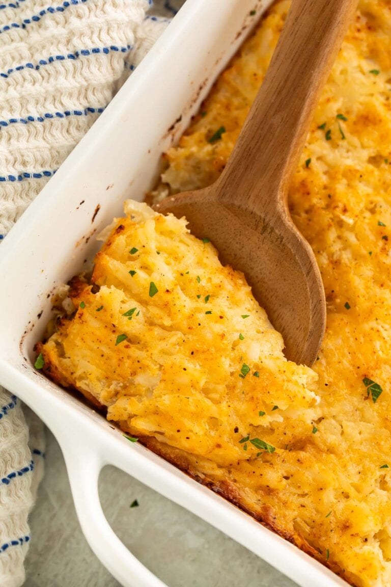 A large wooden spoon resting in a square casserole dish holding a cheesy potato casserole.